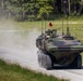 A wheeled armored military vehicle, consistent with a Light Armored Vehicle (LAV) or similar 8-wheeled APC, is traveling at speed along a gravel road kicking up a dust cloud. The vehicle is painted in green camouflage and is equipped with a mounted weapon system and multiple antennas. The setting appears to be a military training area surrounded by pine trees and open grassland.