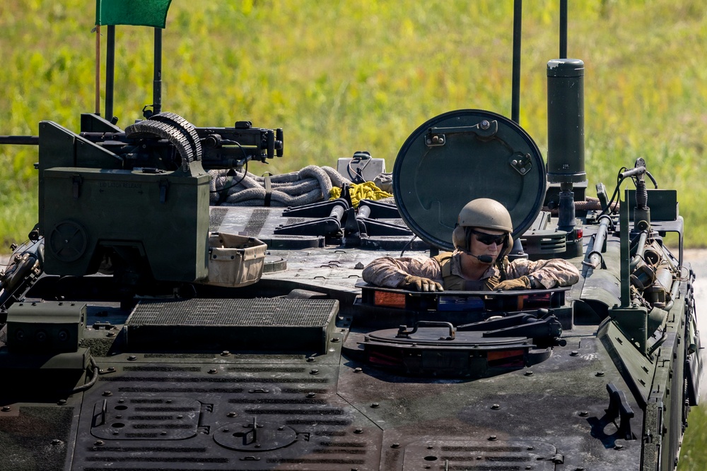 A military crew member is visible from the waist up through an open hatch of an armored fighting vehicle. The soldier is wearing a combat helmet, sunglasses, and camouflage uniform with a communications headset. The vehicle's top deck shows various equipment, tools, cables, and external components consistent with a modern armored personnel carrier or infantry fighting vehicle.