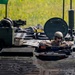 A military crew member is visible from the waist up through an open hatch of an armored fighting vehicle. The soldier is wearing a combat helmet, sunglasses, and camouflage uniform with a communications headset. The vehicle's top deck shows various equipment, tools, cables, and external components consistent with a modern armored personnel carrier or infantry fighting vehicle.
