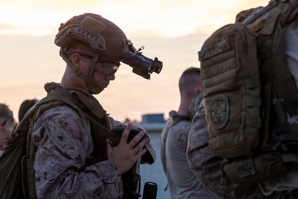 A military service member in full combat gear is pictured at dusk or dawn, appearing to inspect or handle equipment. The soldier is wearing MARPAT desert camouflage, a tactical helmet with a night vision device mount, and full body armor. Additional personnel in similar gear are visible in the background.