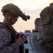 A military service member in full combat gear is pictured at dusk or dawn, appearing to inspect or handle equipment. The soldier is wearing MARPAT desert camouflage, a tactical helmet with a night vision device mount, and full body armor. Additional personnel in similar gear are visible in the background.