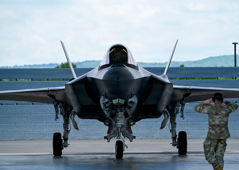 A front-facing view of an F-35 Lightning II stealth fighter jet on a tarmac apron, with its landing gear deployed and weapons bay door open. A military personnel member in camouflage uniform is visible to the right, appearing to salute or shield their eyes. The setting appears to be a military airbase with a blast wall and tree-covered hills visible in the background.