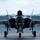 The F-35A Lightning II offically arrives at the 104th Fighter Wing, brings new era to Barnes Air National Guard Base A front-facing view of an F-35 Lightning II stealth fighter jet on a tarmac apron, with its landing gear deployed and weapons bay door open. A military personnel member in camouflage uniform is visible to the right, appearing to salute or shield their eyes. The setting appears to be a military airbase with a blast wall and tree-covered hills visible in the background.