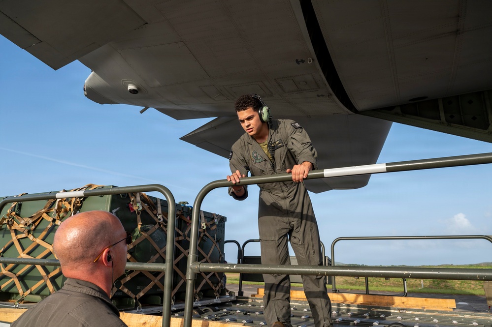 A military personnel member in a flight suit and green hearing protection headset is working on an aircraft loading ramp, handling a metal bar or roller component. A second individual, bald and wearing glasses, is visible from behind in the foreground. Netted cargo pallets with military equipment are staged on the tarmac in the background.