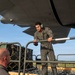 A military personnel member in a flight suit and green hearing protection headset is working on an aircraft loading ramp, handling a metal bar or roller component. A second individual, bald and wearing glasses, is visible from behind in the foreground. Netted cargo pallets with military equipment are staged on the tarmac in the background.