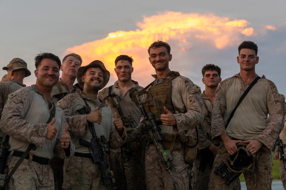 A group of military personnel, appearing to be U.S. Marines based on their desert MARPAT camouflage uniforms, pose together for a group photo at dusk. Several individuals are giving thumbs-up gestures and smiling, suggesting the completion of a successful training exercise or mission. A dramatic illuminated cloud formation is visible in the background sky.