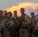 A group of military personnel, appearing to be U.S. Marines based on their desert MARPAT camouflage uniforms, pose together for a group photo at dusk. Several individuals are giving thumbs-up gestures and smiling, suggesting the completion of a successful training exercise or mission. A dramatic illuminated cloud formation is visible in the background sky.