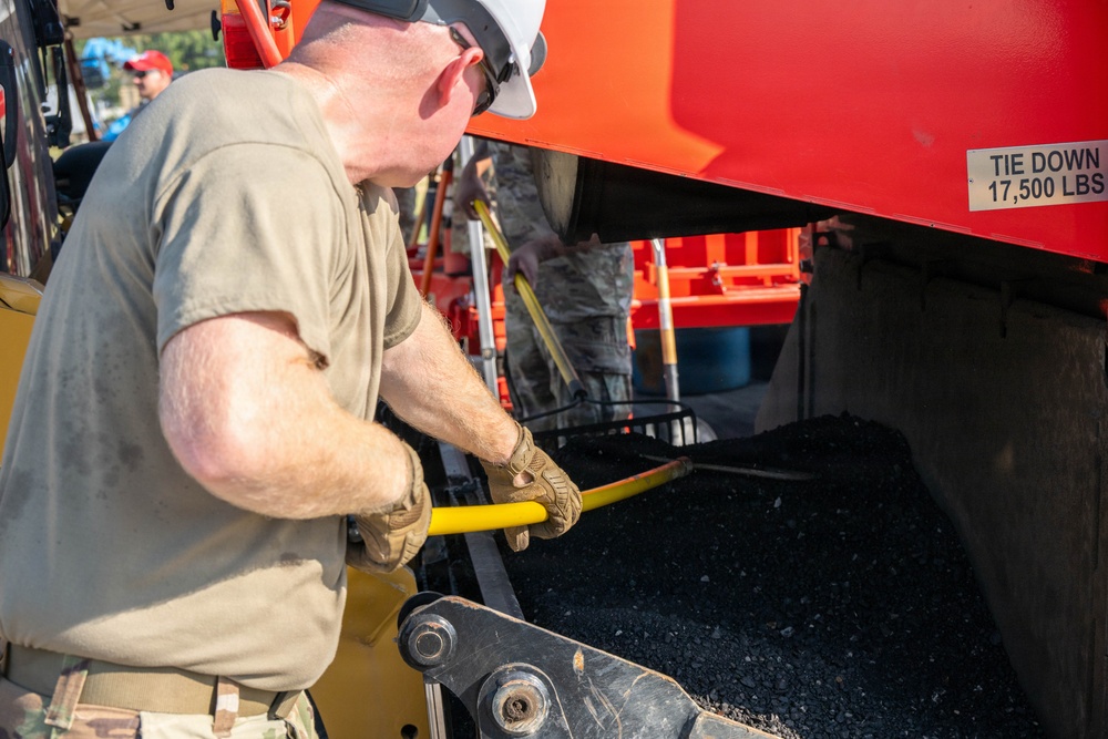 A worker wearing a white hard hat, safety glasses, and tactical gloves is operating or adjusting equipment on a red paving or road construction machine. The machine contains dark asphalt aggregate material and is labeled with a 'TIE DOWN 17,500 LBS' specification plate. At least one additional person in military-style camouflage uniform is visible in the background.