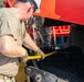 A worker wearing a white hard hat, safety glasses, and tactical gloves is operating or adjusting equipment on a red paving or road construction machine. The machine contains dark asphalt aggregate material and is labeled with a 'TIE DOWN 17,500 LBS' specification plate. At least one additional person in military-style camouflage uniform is visible in the background.