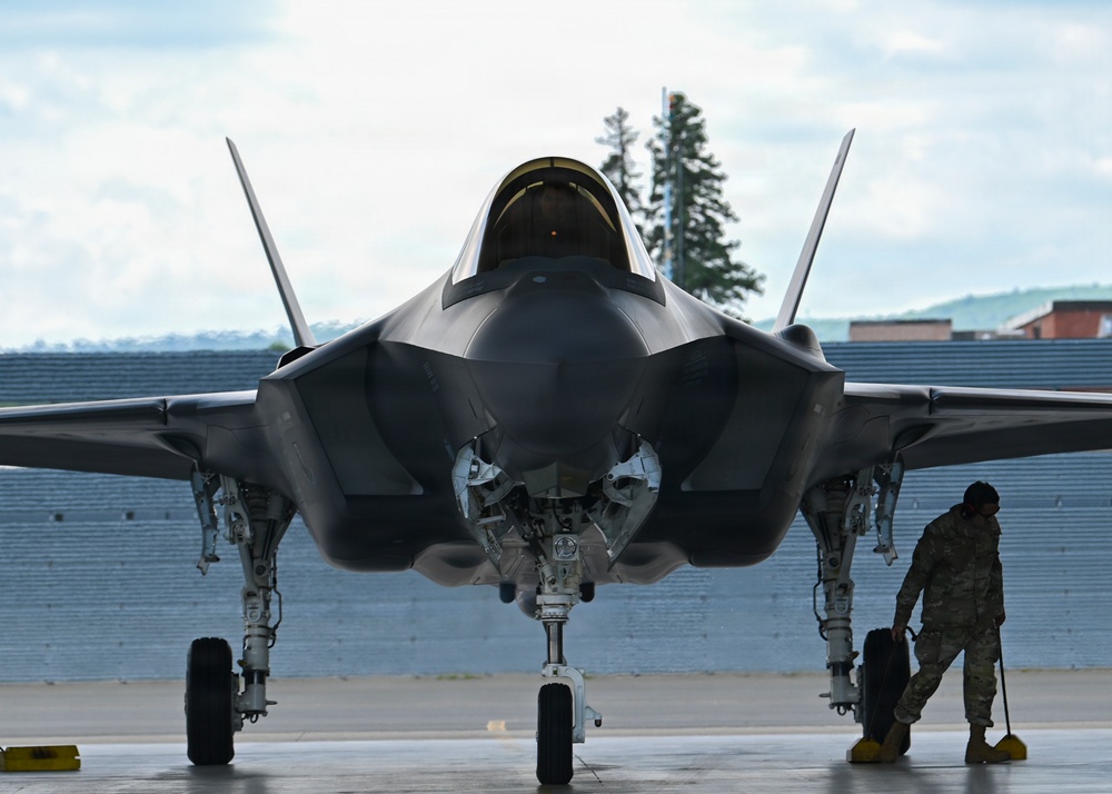 A front-facing view of an F-35 Lightning II stealth fighter jet parked on a flight line apron. A military ground crew member in camouflage uniform is visible to the right side of the aircraft, apparently conducting a pre- or post-flight inspection. The aircraft's nose gear bay is open and wheel chocks are visible beneath the landing gear.