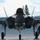 The F-35A Lightning II offically arrives at the 104th Fighter Wing, brings new era to Barnes Air National Guard Base A front-facing view of an F-35 Lightning II stealth fighter jet parked on a flight line apron. A military ground crew member in camouflage uniform is visible to the right side of the aircraft, apparently conducting a pre- or post-flight inspection. The aircraft's nose gear bay is open and wheel chocks are visible beneath the landing gear.