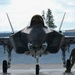 A front-facing view of an F-35 Lightning II stealth fighter jet parked on a flight line apron. A military ground crew member in camouflage uniform is visible to the right side of the aircraft, apparently conducting a pre- or post-flight inspection. The aircraft's nose gear bay is open and wheel chocks are visible beneath the landing gear.