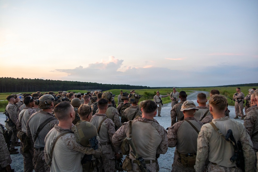 A large group of military personnel, appearing to be U.S. Marines based on their distinctive MARPAT camouflage uniforms, are gathered in a semicircle on a paved road or airstrip during twilight hours. One or two individuals at the front appear to be addressing the group, suggesting a pre-mission briefing or after-action review. The setting appears to be a rural training area with open fields and tree lines visible in the background.