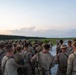 A large group of military personnel, appearing to be U.S. Marines based on their distinctive MARPAT camouflage uniforms, are gathered in a semicircle on a paved road or airstrip during twilight hours. One or two individuals at the front appear to be addressing the group, suggesting a pre-mission briefing or after-action review. The setting appears to be a rural training area with open fields and tree lines visible in the background.