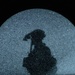 The image shows a dark silhouette of a human figure cast onto a large spherical object with a grainy, glittery teal-gray surface. The sphere is set against a completely black background, creating a stark contrast. The silhouette appears to depict a person, possibly holding an object or wearing a hat, rendered as a shadow projection on the sphere's surface.