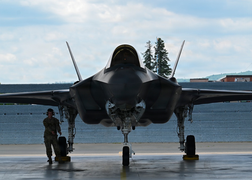 A front-facing view of an F-35 Lightning II stealth fighter jet parked on a tarmac apron, with landing gear deployed and canopy closed. A single military ground crew member in camouflage uniform stands to the left of the aircraft wearing hearing protection. The setting appears to be a military air base with a brick wall structure and trees visible in the background.