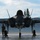 The F-35A Lightning II offically arrives at the 104th Fighter Wing, brings new era to Barnes Air National Guard Base A front-facing view of an F-35 Lightning II stealth fighter jet parked on a tarmac apron, with landing gear deployed and canopy closed. A single military ground crew member in camouflage uniform stands to the left of the aircraft wearing hearing protection. The setting appears to be a military air base with a brick wall structure and trees visible in the background.
