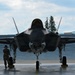 A front-facing view of an F-35 Lightning II stealth fighter jet parked on a tarmac apron, with landing gear deployed and canopy closed. A single military ground crew member in camouflage uniform stands to the left of the aircraft wearing hearing protection. The setting appears to be a military air base with a brick wall structure and trees visible in the background.