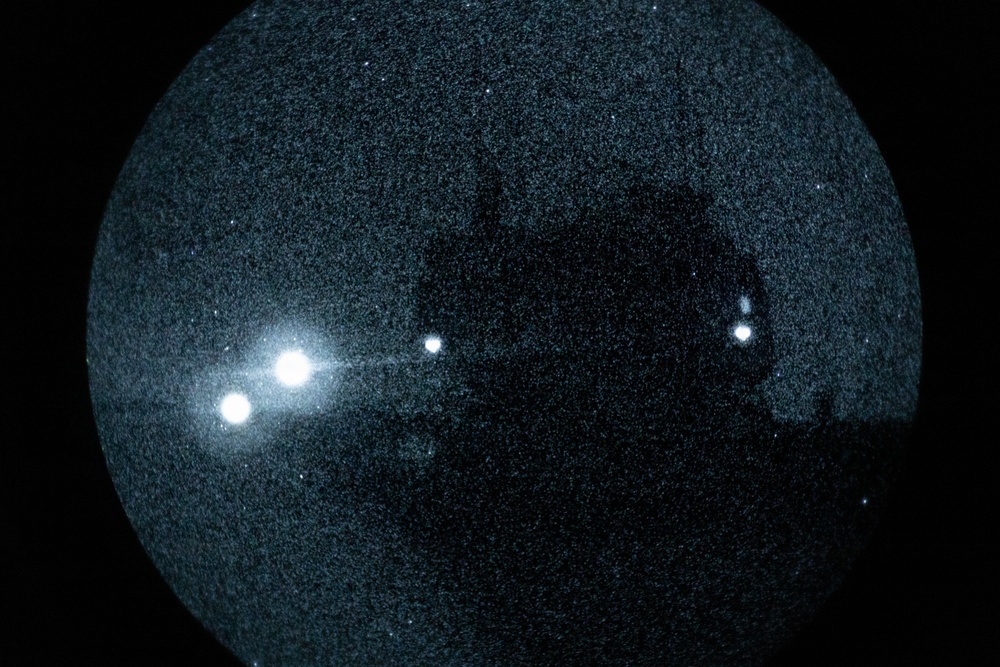 An all-sky or fisheye lens photograph of the night sky, captured in a circular frame against a black background. The image shows several bright point sources of light distributed across the field, with a dark silhouette of what appears to be a structure or tree visible in the upper-right quadrant. The image has significant grain/noise, consistent with a long-exposure or low-light night sky camera.
