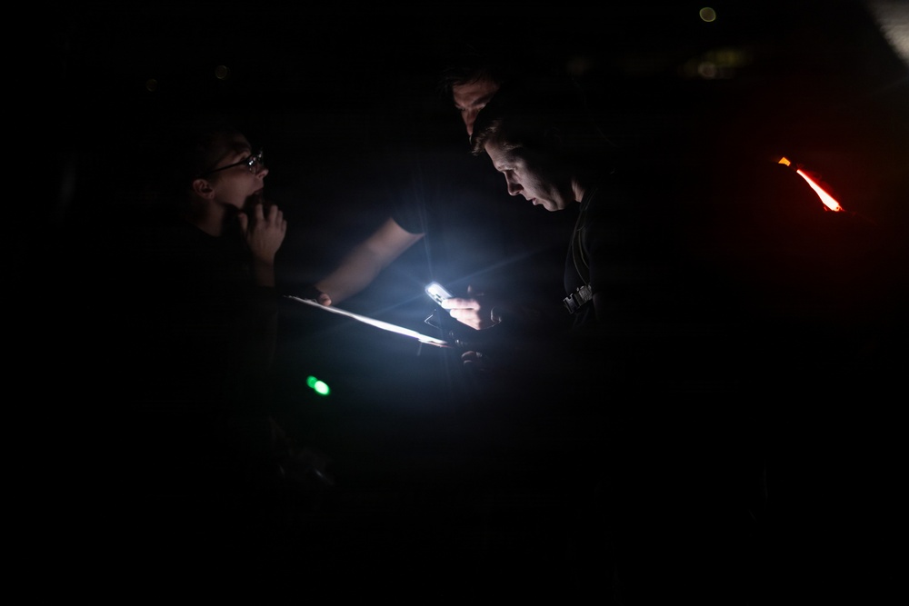 Three individuals are gathered in a very dark environment, illuminated primarily by the glow of a smartphone and what appears to be a tablet or clipboard. The central figure is using a phone as a light source while examining a document or device. Small colored indicator lights (green and red) are visible in the background.