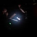 Three individuals are gathered in a very dark environment, illuminated primarily by the glow of a smartphone and what appears to be a tablet or clipboard. The central figure is using a phone as a light source while examining a document or device. Small colored indicator lights (green and red) are visible in the background.