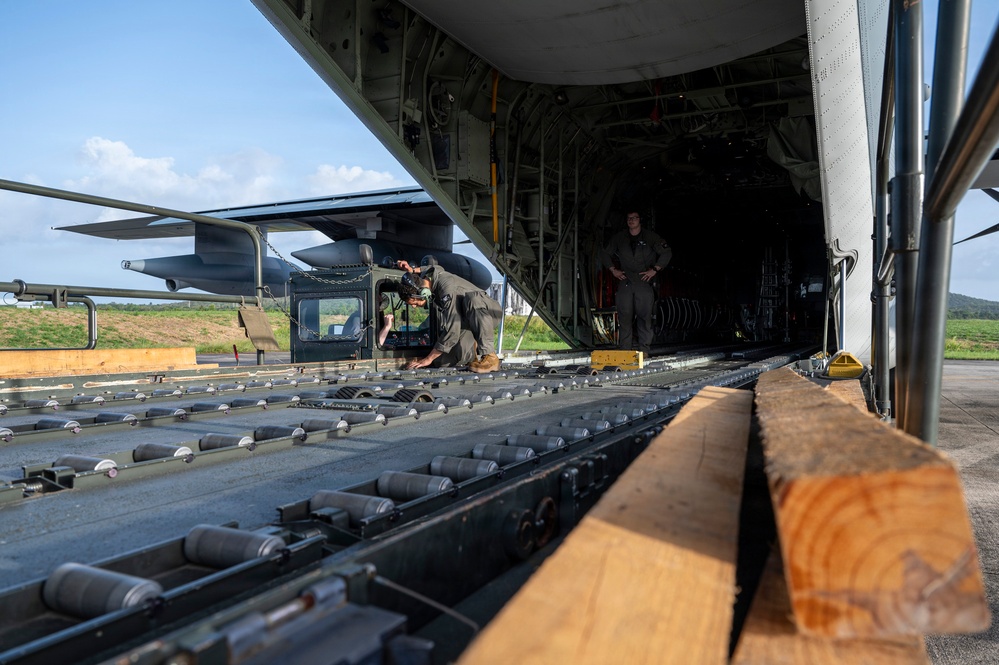 Military personnel are loading or securing a helicopter onto a large military cargo aircraft, likely a C-17 or C-130 variant, via its rear ramp. The ramp features roller conveyors and tie-down tracks, with wooden planks visible as loading aids. Two personnel are visible — one crouching and working near the helicopter, another standing inside the cargo bay observing.