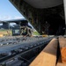 Military personnel are loading or securing a helicopter onto a large military cargo aircraft, likely a C-17 or C-130 variant, via its rear ramp. The ramp features roller conveyors and tie-down tracks, with wooden planks visible as loading aids. Two personnel are visible — one crouching and working near the helicopter, another standing inside the cargo bay observing.