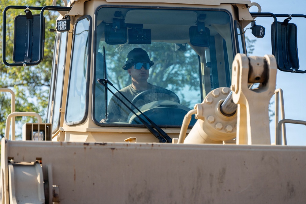 A male operator is seated inside the cab of a large tan/beige heavy construction vehicle, likely a bulldozer or wheel loader. He is wearing blue reflective sunglasses and a dark bucket hat, and appears to be actively operating the machine. The image is taken from a low front angle, emphasizing the vehicle's cab and hydraulic components.