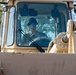 A male operator is seated inside the cab of a large tan/beige heavy construction vehicle, likely a bulldozer or wheel loader. He is wearing blue reflective sunglasses and a dark bucket hat, and appears to be actively operating the machine. The image is taken from a low front angle, emphasizing the vehicle's cab and hydraulic components.