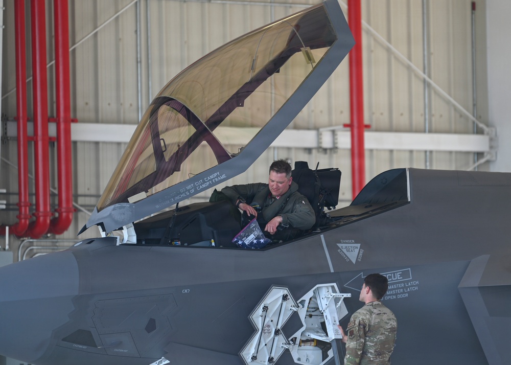 A military pilot in a green flight suit sits in the open cockpit of an F-35 stealth fighter jet inside a hangar. The pilot appears to be interacting with a ground crew member standing beside the aircraft. A folded American flag is visible in the cockpit, suggesting a ceremonial or commemorative occasion.