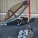A military pilot in a green flight suit sits in the open cockpit of an F-35 stealth fighter jet inside a hangar. The pilot appears to be interacting with a ground crew member standing beside the aircraft. A folded American flag is visible in the cockpit, suggesting a ceremonial or commemorative occasion.
