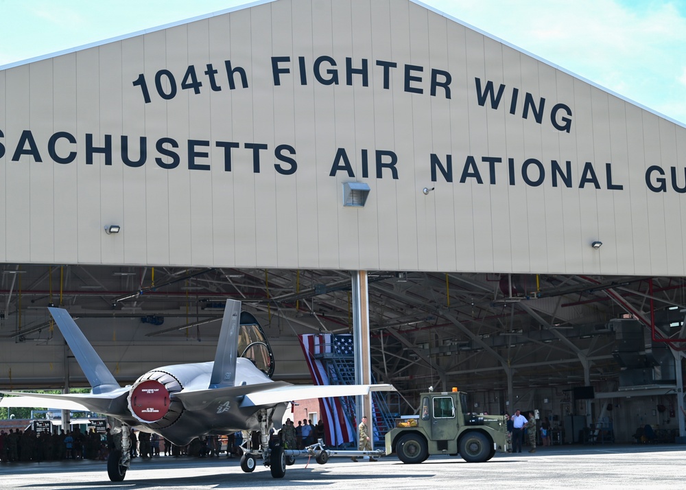 An F-35 fighter jet is being towed out of a large military hangar belonging to the 104th Fighter Wing, Massachusetts Air National Guard. The scene appears to be a ceremonial or official event, evidenced by the gathered personnel, American flags displayed inside the hangar, and the formal presentation of the aircraft. A ground support tow vehicle is attached to the front landing gear of the aircraft.