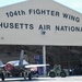 An F-35 fighter jet is being towed out of a large military hangar belonging to the 104th Fighter Wing, Massachusetts Air National Guard. The scene appears to be a ceremonial or official event, evidenced by the gathered personnel, American flags displayed inside the hangar, and the formal presentation of the aircraft. A ground support tow vehicle is attached to the front landing gear of the aircraft.