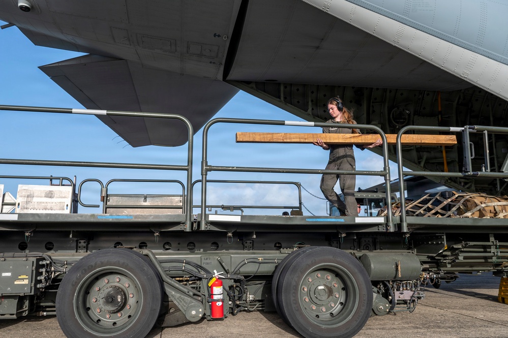 A ground crew member wearing headphones is working on a military cargo loader positioned beneath the wing of a large military transport aircraft. The worker is handling a wooden plank or board, likely used as a loading aid or shim during cargo operations. A red fire extinguisher is mounted on the loader vehicle as a safety precaution.