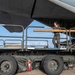 A ground crew member wearing headphones is working on a military cargo loader positioned beneath the wing of a large military transport aircraft. The worker is handling a wooden plank or board, likely used as a loading aid or shim during cargo operations. A red fire extinguisher is mounted on the loader vehicle as a safety precaution.