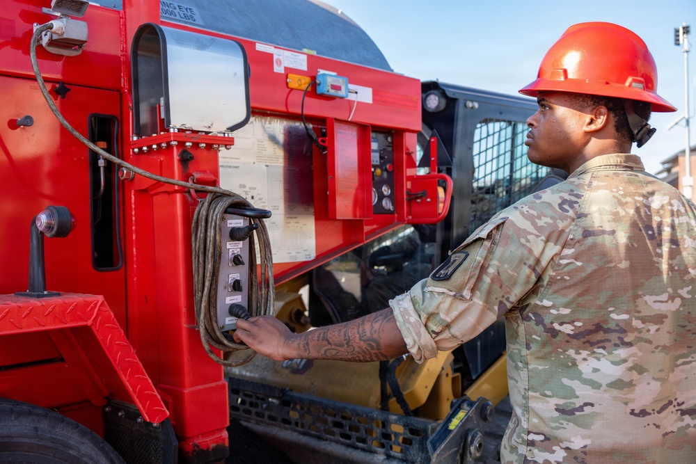 A military service member wearing OCP (Operational Camouflage Pattern) uniform and a red hard hat is operating a control panel on a large red piece of heavy equipment or vehicle. The individual appears to be performing maintenance or operational checks on the machinery. The setting appears to be an outdoor military or construction work site.
