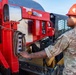 A military service member wearing OCP (Operational Camouflage Pattern) uniform and a red hard hat is operating a control panel on a large red piece of heavy equipment or vehicle. The individual appears to be performing maintenance or operational checks on the machinery. The setting appears to be an outdoor military or construction work site.