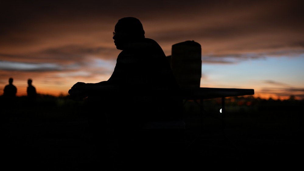 A silhouetted figure sits or leans against what appears to be a chair or bench, set against a dramatic sunset sky with deep orange, red, and blue tones. The scene is captured in near-total darkness with the sky providing the primary light source. Two additional silhouetted figures are faintly visible in the background to the left.