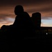 A silhouetted figure sits or leans against what appears to be a chair or bench, set against a dramatic sunset sky with deep orange, red, and blue tones. The scene is captured in near-total darkness with the sky providing the primary light source. Two additional silhouetted figures are faintly visible in the background to the left.