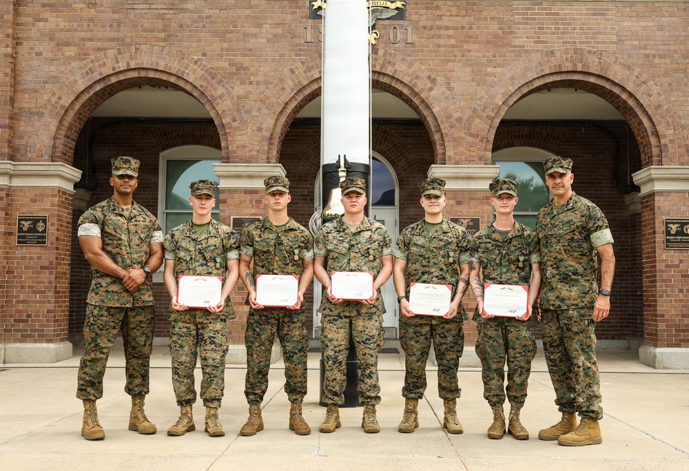 Seven U.S. Marines in woodland MARPAT camouflage uniforms are posed in a group photo in front of a red brick building with arched entrances. Five of the Marines in the center are holding red-bordered certificates, suggesting an awards or recognition ceremony. Two Marines on the far left and far right appear to be senior personnel presenting or overseeing the ceremony.