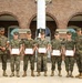Seven U.S. Marines in woodland MARPAT camouflage uniforms are posed in a group photo in front of a red brick building with arched entrances. Five of the Marines in the center are holding red-bordered certificates, suggesting an awards or recognition ceremony. Two Marines on the far left and far right appear to be senior personnel presenting or overseeing the ceremony.