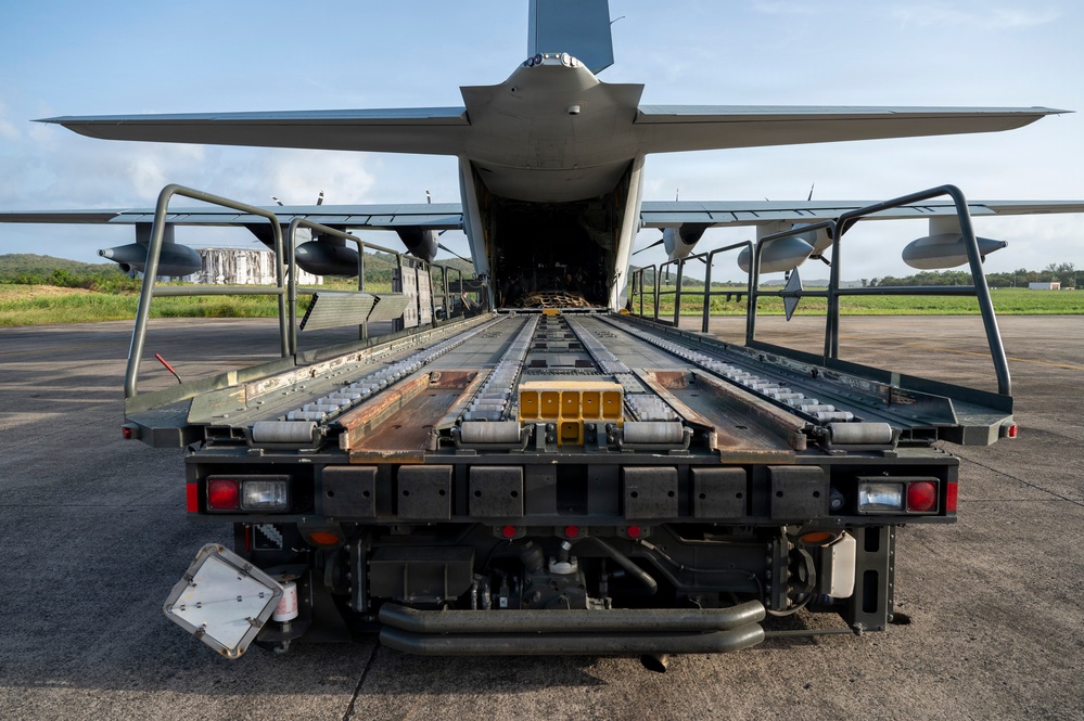 A military cargo aircraft, likely a C-130 Hercules or similar transport, is shown with its rear cargo ramp open and aligned with a flatbed cargo loader vehicle. The perspective is from directly behind the loader, looking straight into the aircraft's cargo hold. The scene appears to be at a military airfield or forward operating base, suggesting active loading or unloading operations.