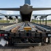 A military cargo aircraft, likely a C-130 Hercules or similar transport, is shown with its rear cargo ramp open and aligned with a flatbed cargo loader vehicle. The perspective is from directly behind the loader, looking straight into the aircraft's cargo hold. The scene appears to be at a military airfield or forward operating base, suggesting active loading or unloading operations.