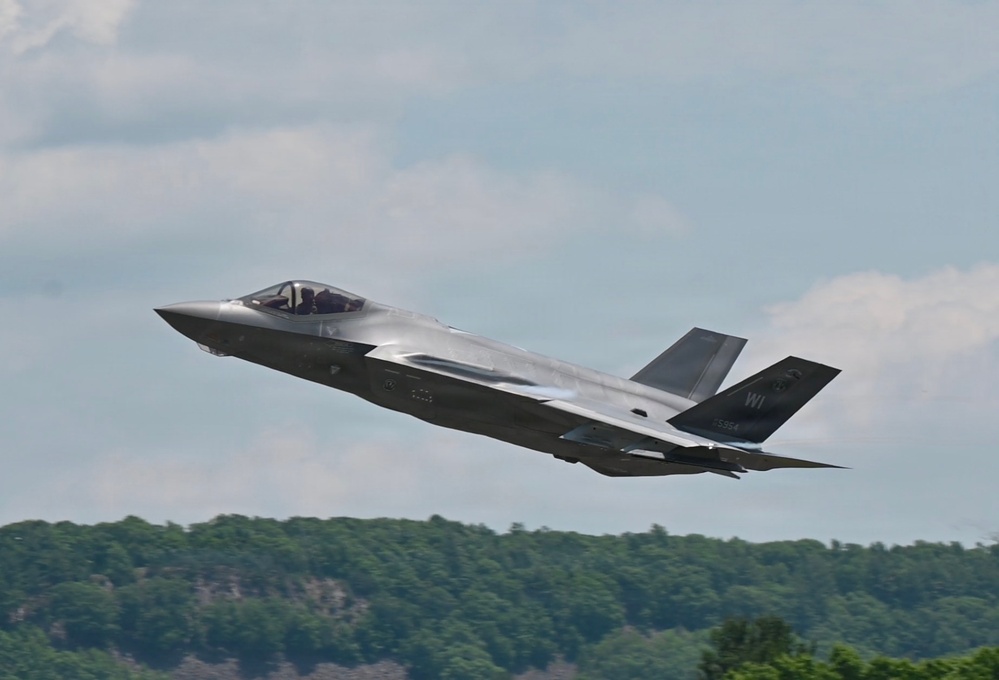 A single-seat stealth fighter jet, identifiable as an F-35A Lightning II, is captured in low-altitude flight just after takeoff or during a low pass. The aircraft bears tail markings including the letters 'WI' and a partial serial number visible on the vertical stabilizer, suggesting Wisconsin Air National Guard affiliation. The background features a dense treeline and partly cloudy sky.