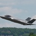 A single-seat stealth fighter jet, identifiable as an F-35A Lightning II, is captured in low-altitude flight just after takeoff or during a low pass. The aircraft bears tail markings including the letters 'WI' and a partial serial number visible on the vertical stabilizer, suggesting Wisconsin Air National Guard affiliation. The background features a dense treeline and partly cloudy sky.