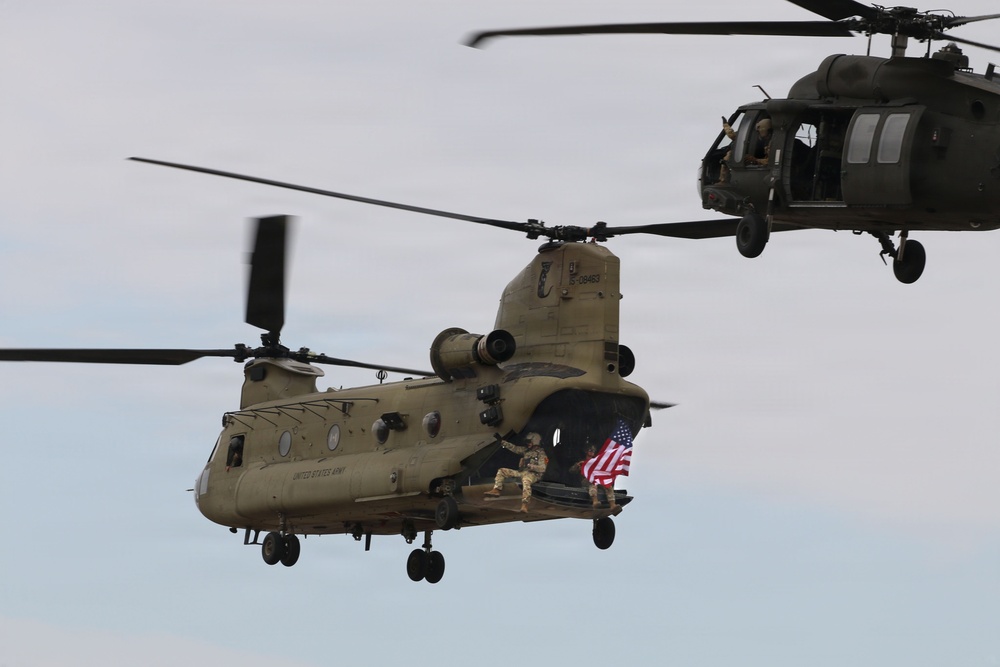 Two U.S. Army helicopters are flying in formation against an overcast sky. The primary aircraft is a CH-47 Chinook tandem-rotor helicopter marked 'UNITED STATES ARMY,' with soldiers visible at the rear ramp displaying an American flag. A UH-60 Black Hawk helicopter flies in the background to the upper right.