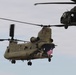 Two U.S. Army helicopters are flying in formation against an overcast sky. The primary aircraft is a CH-47 Chinook tandem-rotor helicopter marked 'UNITED STATES ARMY,' with soldiers visible at the rear ramp displaying an American flag. A UH-60 Black Hawk helicopter flies in the background to the upper right.