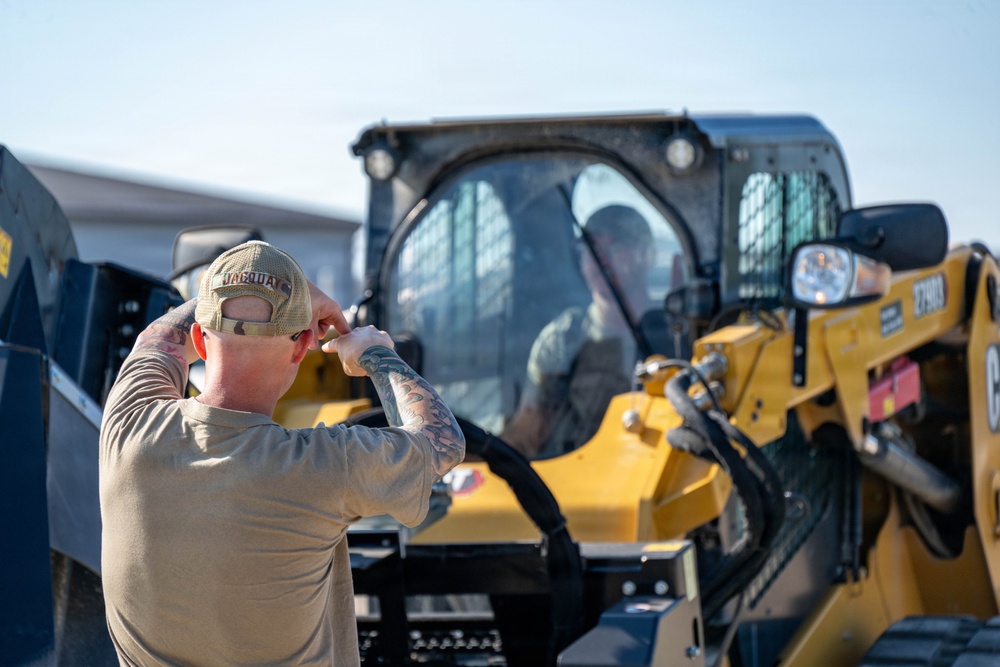 A tattooed man wearing a tan cap and khaki shirt is directing or signaling to an operator inside a yellow skid steer loader (appears to be a Caterpillar model). The ground guide is using hand signals to communicate with the equipment operator. The scene appears to be an outdoor worksite or staging area.