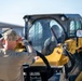 A tattooed man wearing a tan cap and khaki shirt is directing or signaling to an operator inside a yellow skid steer loader (appears to be a Caterpillar model). The ground guide is using hand signals to communicate with the equipment operator. The scene appears to be an outdoor worksite or staging area.