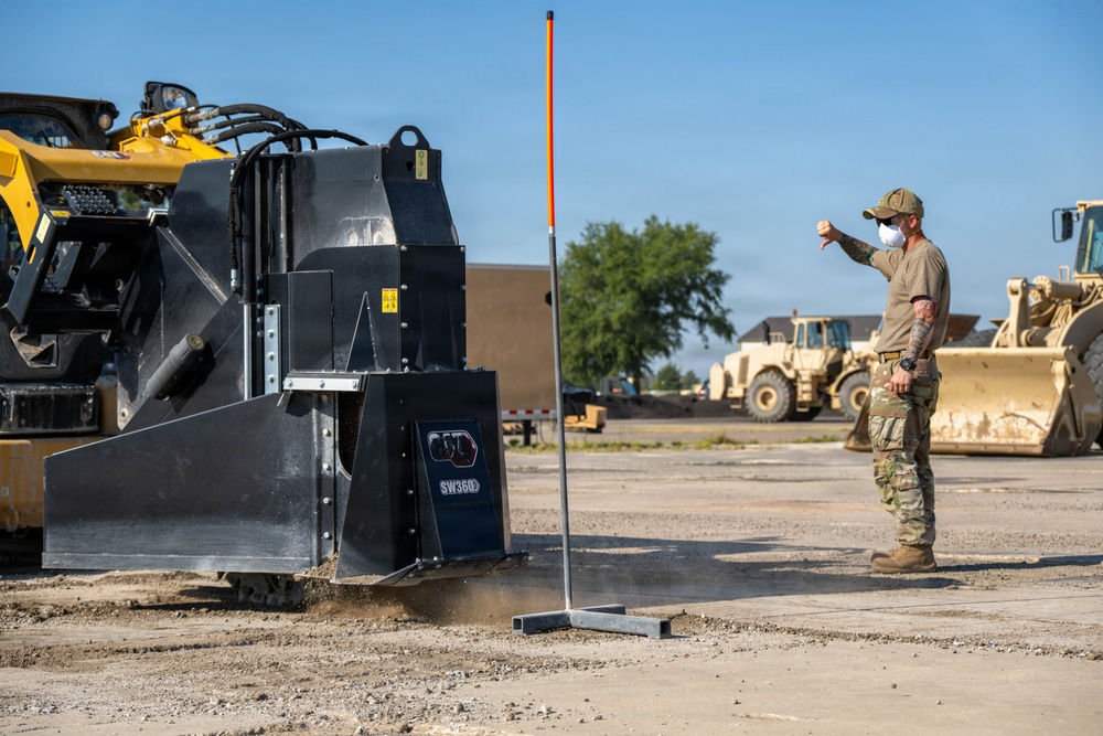 A person in military-style camouflage clothing and a face mask is giving a thumbs-down hand signal near heavy construction equipment on a gravel surface. The scene appears to be a military or construction staging area with multiple pieces of heavy machinery visible. An orange-tipped marker pole is positioned between the worker and the equipment.
